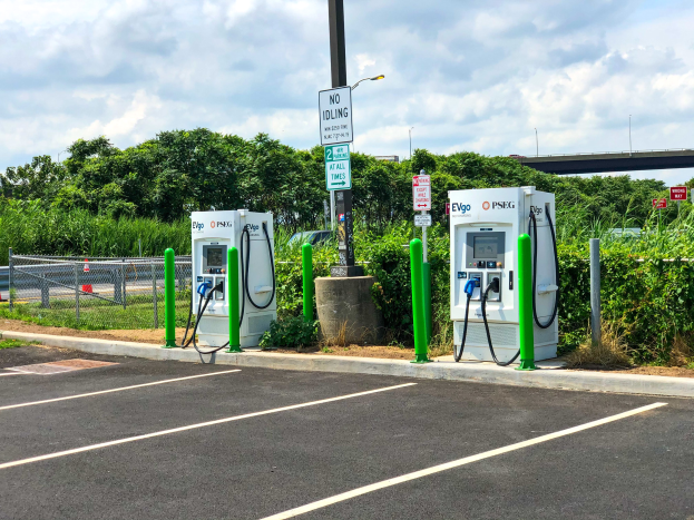 Elektrofahrzeug-Ladestation auf einem Parkplatz mit umgebender Infrastruktur, Vegetation und einem bewölkten Himmel.