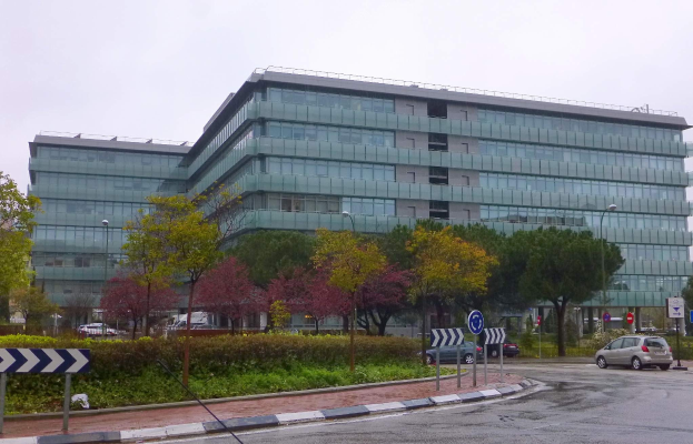 Großes modernes Gebäude mit zahlreichen Fenstern, identifiziert als Hauptsitz der Europäischen Zentralbank, umgeben von Grünflächen, Straßeninfrastruktur und Fahrzeugen unter einem bewölkten Himmel.
