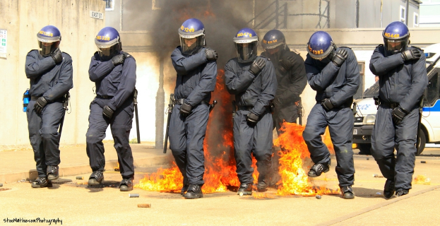 Männer mit Helmen stehen vor einem Feuer mit verschiedenen Gegenständen auf dem Boden, Gebäuden im Hintergrund, einem Fahrzeug und einem Plakat und einer Tafel an der linken Wand; Text ist am unteren Bildrand sichtbar.