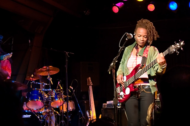Eine Frau in einer grünen Jacke spielt Gitarre auf der Bühne während eines Konzerts, mit einer Bandaufstellung zu ihrer Linken und Lichtern oben in einem dunklen Hintergrund.