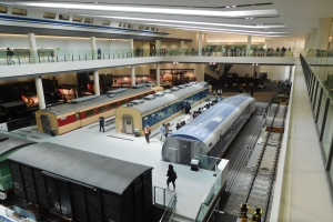 Ein geräumiger Museumsraum voller verschiedener Züge auf Display, Menschen beim Beobachten, Geländer, Säulen und Deckenbeleuchtung.