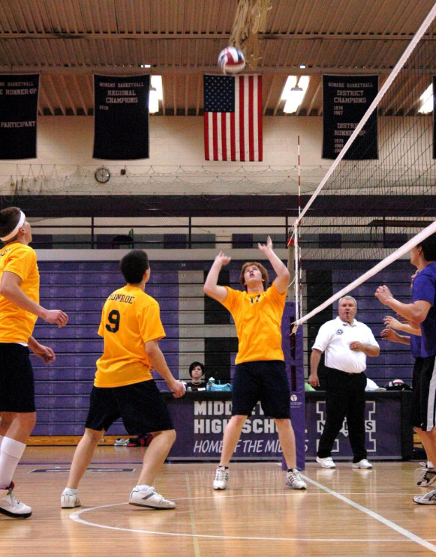 Menschengruppen auf einem Spielfeld mit Netz und Ball, bekleidet und beschuht, mit Landesflagge, Wand und Plakaten mit Text.