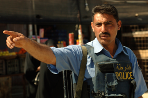 Ein Polizeibeamter in Uniform zeigt mit einer Waffe in der Hand auf etwas, vor einem Laden mit unscharfen Menschen und Gegenst├Ąnden im Hintergrund.