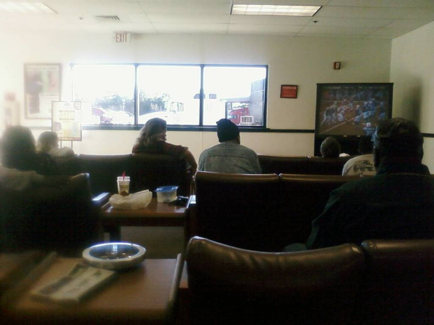 Ein Raum mit einer Decke, einem Fenster, einer Couch mit sitzenden Menschen, einem Tisch mit einer Tasse, Schachtel, Abdeckung, Buch und Schale, einem Banner an der Wand, einem Mann mit M├╝tze und einem Ausgangsschild.