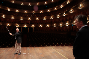Ein älterer Mann in einem Anzug hält einen Golfschläger auf der Bühne, während ein weiterer älterer Mann rechts von ihm steht, unter Deckenlampen mit vielen Sitzplätzen darunter.
