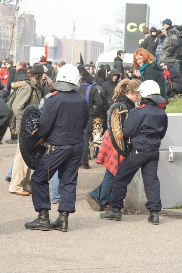 Eine Gruppe von Menschen, die auf einer Straße gehen, mit zwei Personen in der Vorderreihe, die wie Polizisten aussehen, Gebäuden im Hintergrund und Boden unten.
