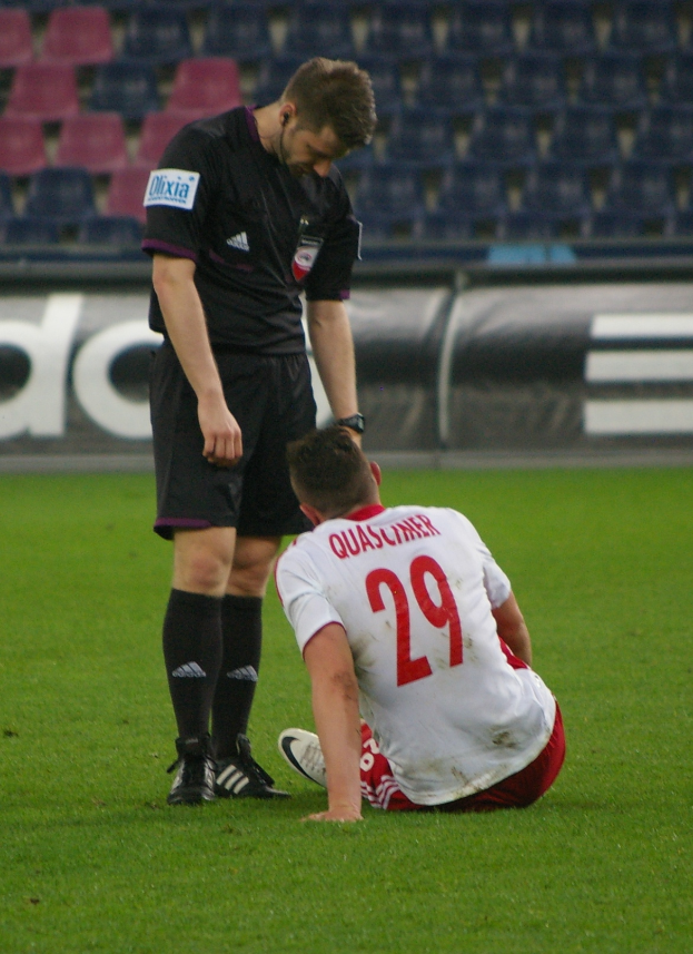 Ein Fußballspieler sitzt neben einem Schiedsrichter auf dem Boden in einem Stadion, beide tragen Sportkleidung, mit Plakaten und Stühlen im Hintergrund.