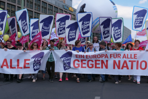 Eine Gruppe von Menschen marschiert mit einem Banner, auf dem 'Ein Europa für alle gegen Nation' steht, und verschiedenen Plakaten die Straße entlang. Im Hintergrund sind Gebäude, Luftballons und ein klarer blauer Himmel zu sehen.