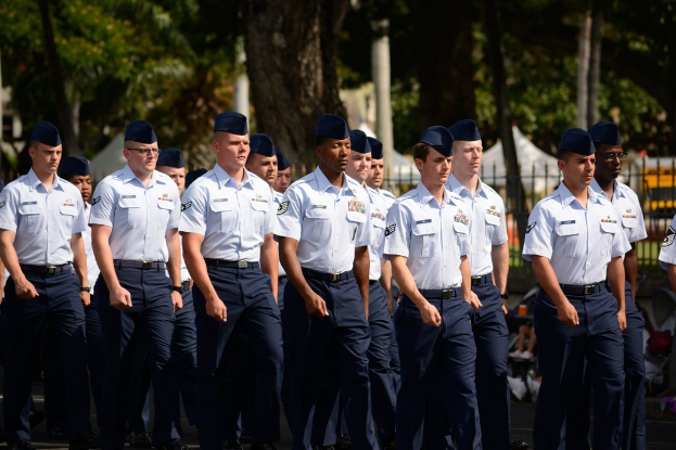 Männer in Uniform auf einer Straße mit einem Eisenzaun dahinter, Bäume und Pfosten im Hintergrund.