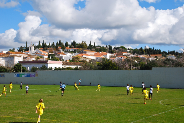 Eine Gruppe von Menschen, die ein Spiel auf einem Feld mit Plakaten an einer nahen Wand, Bäumen, Pfählen, Häusern und Wolken im Hintergrund spielen