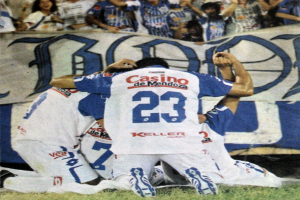 Gruppe von Fußballspielern feiert auf dem Boden vor einer jubelnden Menge, mit einem Banner mit Text im Hintergrund.