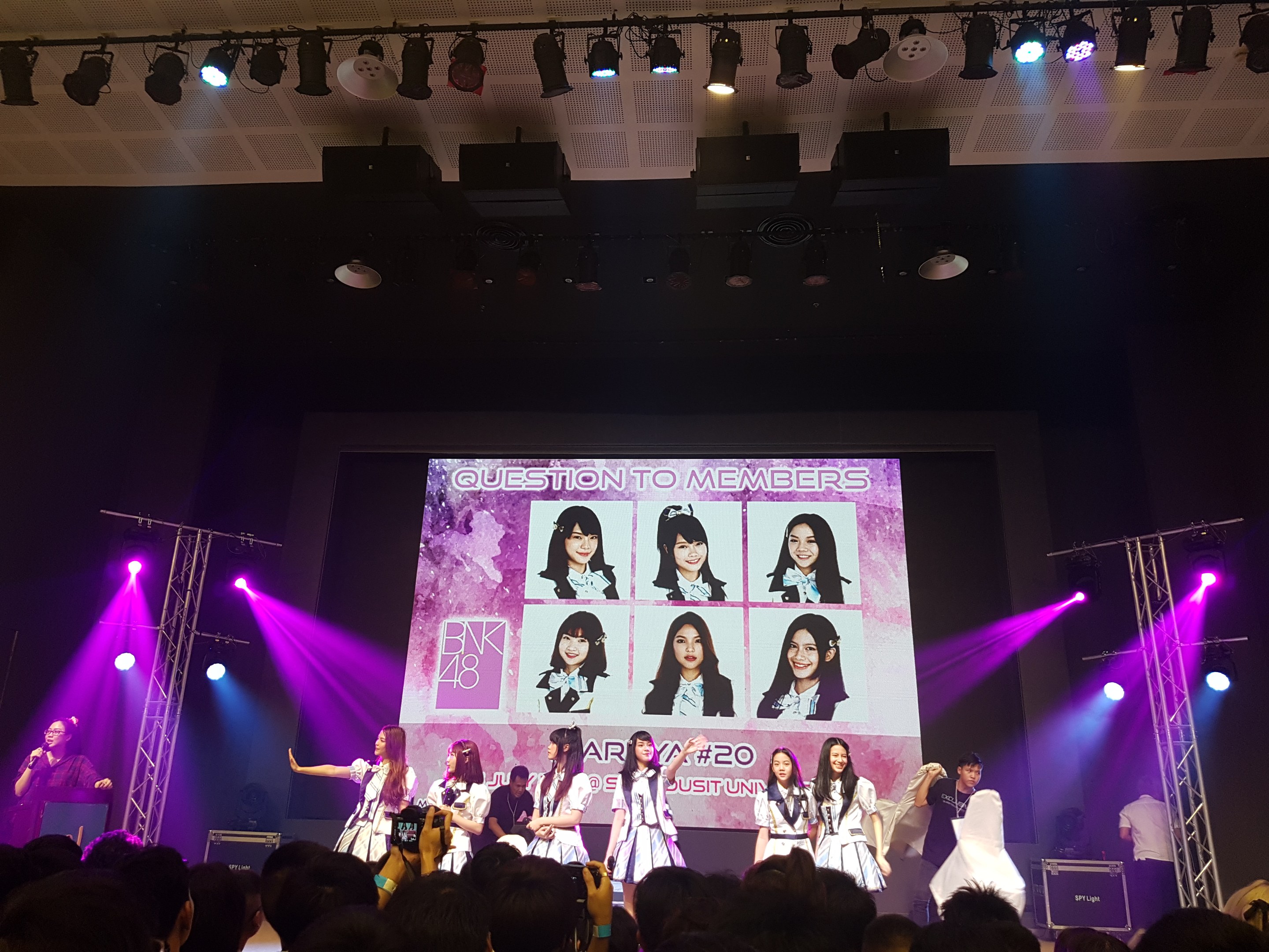 Eine Gruppe von Menschen, die auf einer hell erleuchteten Bühne performs, mit einer Menge darunter, mit einem Sänger mit Mikrofon und einem Musiker, der ein Instrument spielt, während ein großer Bildschirm Bilder der K-Pop-Gruppe BK48 anzeigt.