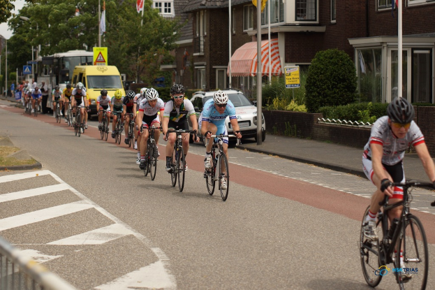 Menschen auf Fahrrädern mit Helmen, parkende Autos auf der Straße und Fahnen an Masten, mit Bäumen und Gebäuden im Hintergrund.