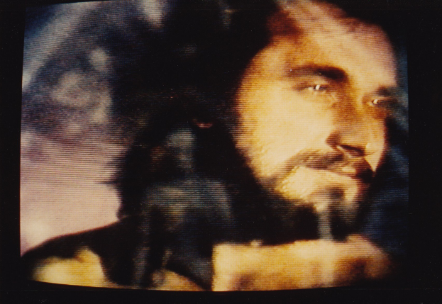 Ein Mann mit langem dunklem Bart und Haar, der ein weißes Hemd trägt, erscheint auf einem Fernsehbildschirm mit ernster Miene und direktem Blick.