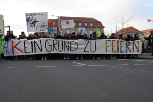 Demonstranten halten ein Transparent mit der Aufschrift "Kein Grund zu Feiern" gegen deutsche Sparmaßnahmen, mit Gebäuden, Bäumen und einem klaren Himmel im Hintergrund.