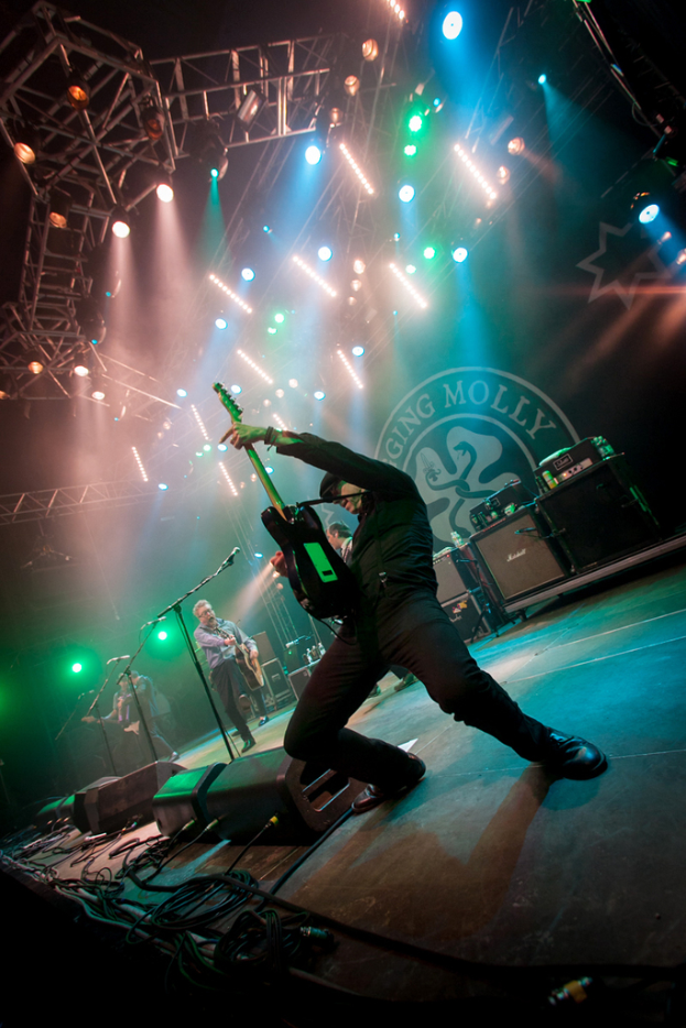 Ein musikalisches Konzert mit drei Könpern auf der Bühne: zwei Gitarristen rechts mit Mikrofonen davor, ein Hintergrund mit 'MOLLEY' und zwei Schlangenlogos sowie Scheinwerfer oben.