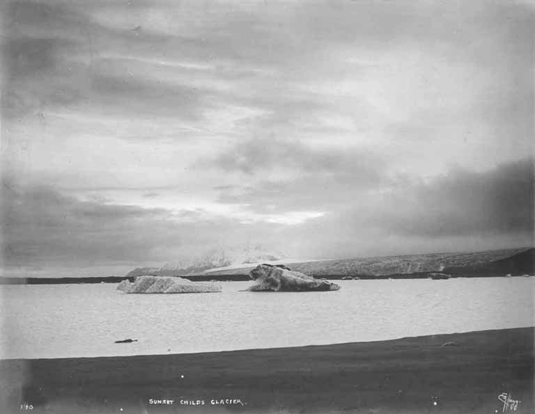 Ein Schwarz-Weiß-Foto eines Eisbergs, der im Ozean treibt, umgeben von Höhen und einem bewölktem Himmel, mit dem Text 'Sunset Childs Glacier' unten.