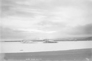 Ein Schwarz-Weiß-Foto eines Eisbergs, der im Ozean treibt, umgeben von Höhen und einem bewölktem Himmel, mit dem Text 'Sunset Childs Glacier' unten.