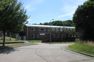 Ein Schulgebäude mit Fenstern befindet sich in der Mitte eines Parks, umgeben von saftigem Grün, mit einem Straßenschild, einem Schild, einer Gruppe von Bäumen, einem Zaun und einem bewölkten Himmel im Hintergrund.