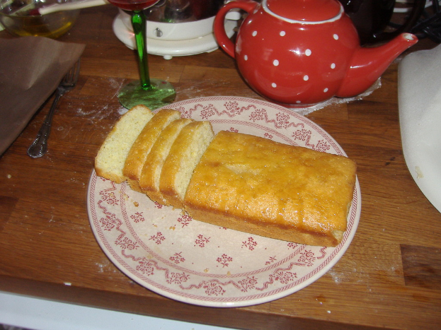 Ein weißer Teller mit Brot auf einer braunen Oberfläche, umgeben von verschiedenen Küchenutensilien, einschließlich einem Glas, einem Kessel, einer Gabel, einem Tablett, einer Schüssel und einem nicht identifizierten weißen Objekt.