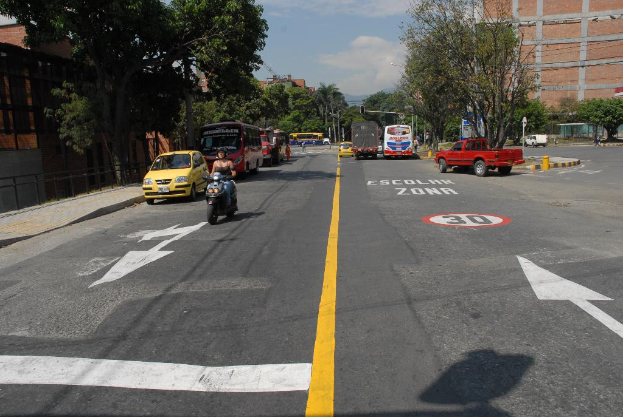 Eine Straßenszene mit Gebäuden, Bäumen, fahrenden Fahrzeugen, Schildern, Verkehrsampeln und Warnschildern auf der Straße unter einem blauen bewölkten Himmel.