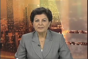 Eine Frau in einem Businessanzug sitzt vor einem Fernseher, mit ein paar Gebäuden im Hintergrund, die als Nachrichtensprecherin mit Text am unteren Bildschirmrand erscheint.