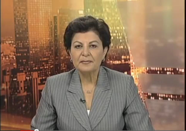 Eine Frau in einem Businessanzug sitzt vor einem Fernseher, mit ein paar Gebäuden im Hintergrund, die als Nachrichtensprecherin mit Text am unteren Bildschirmrand erscheint.