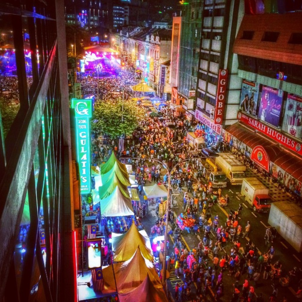 Eine volle KarnevalsSzene mit Zelten, Geschäften, Gebäuden und Bannern, mit beleuchteter Fläche im Hintergrund.