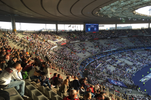 Eine große Menge Menschen in einem Stadion schaut ein Fußballspiel zu, mit einer Bühne rechts, Fahnen, Stangen, einem Bildschirm und der Allianz Arena in München.