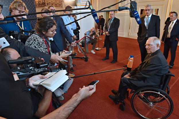 Ein Mann im Rollstuhl spricht mit Reportern, die Kameras, Handys, Mikrofone und eine Fahne mit Text im Hintergrund halten.