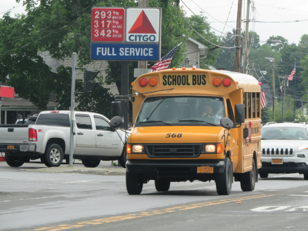 Ein Schulbus fährt auf einer Straße mit sichtbaren Schildern, Strommasten, Kabeln, Straßenlaternen, Ampeln, Gebäuden, Bäumen und einem klaren Himmel.