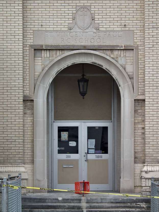 Eingang zu einem Backsteingebäude mit gelbem Absperrband, Treppe, die zu einer Tür mit Papieren daran führt, Maschendrahtzaun auf beiden Seiten und eine hängende Lampe, mit Text an der Wand, der wahrscheinlich den Namen der Schule angibt.