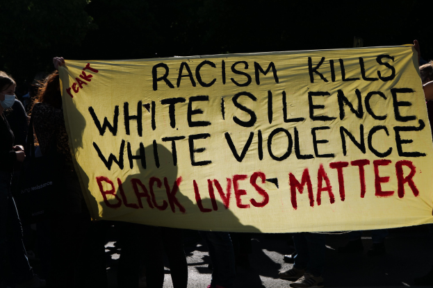 Eine Gruppe maskierter Menschen steht nachts auf einer Straße und hält ein Banner mit der Aufschrift "Rassismus tötet, weiße Stille, weiße Gewalt, Black Lives Matter."