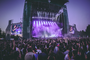 Eine große Menschenmenge bei einem Musikfestival schaut auf der hell erleuchteten Bühne mit Instrumenten und Bildschirmen, die vor einem Gebäude, Bäumen und einem klaren blauen Himmel stehen.