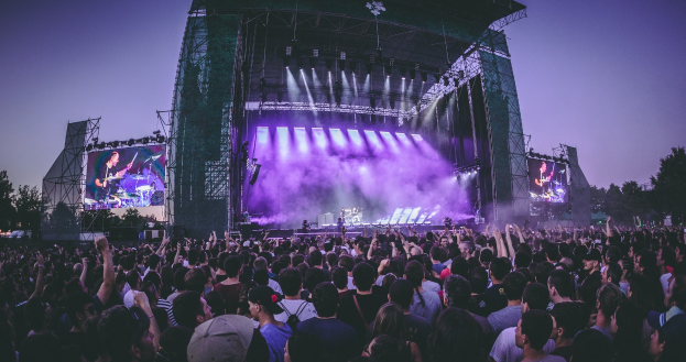 Eine große Menschenmenge bei einem Musikfestival schaut auf der hell erleuchteten Bühne mit Instrumenten und Bildschirmen, die vor einem Gebäude, Bäumen und einem klaren blauen Himmel stehen.