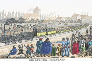 Gruppe von Menschen um einen frühen Zug an Ludwigs-Eisenbahn, Deutschlands erstem Bahnhof, mit sitzenden Passagieren im Inneren und stehenden Individuen außerhalb, vor einem Hintergrund von Gebäuden und Bäumen.