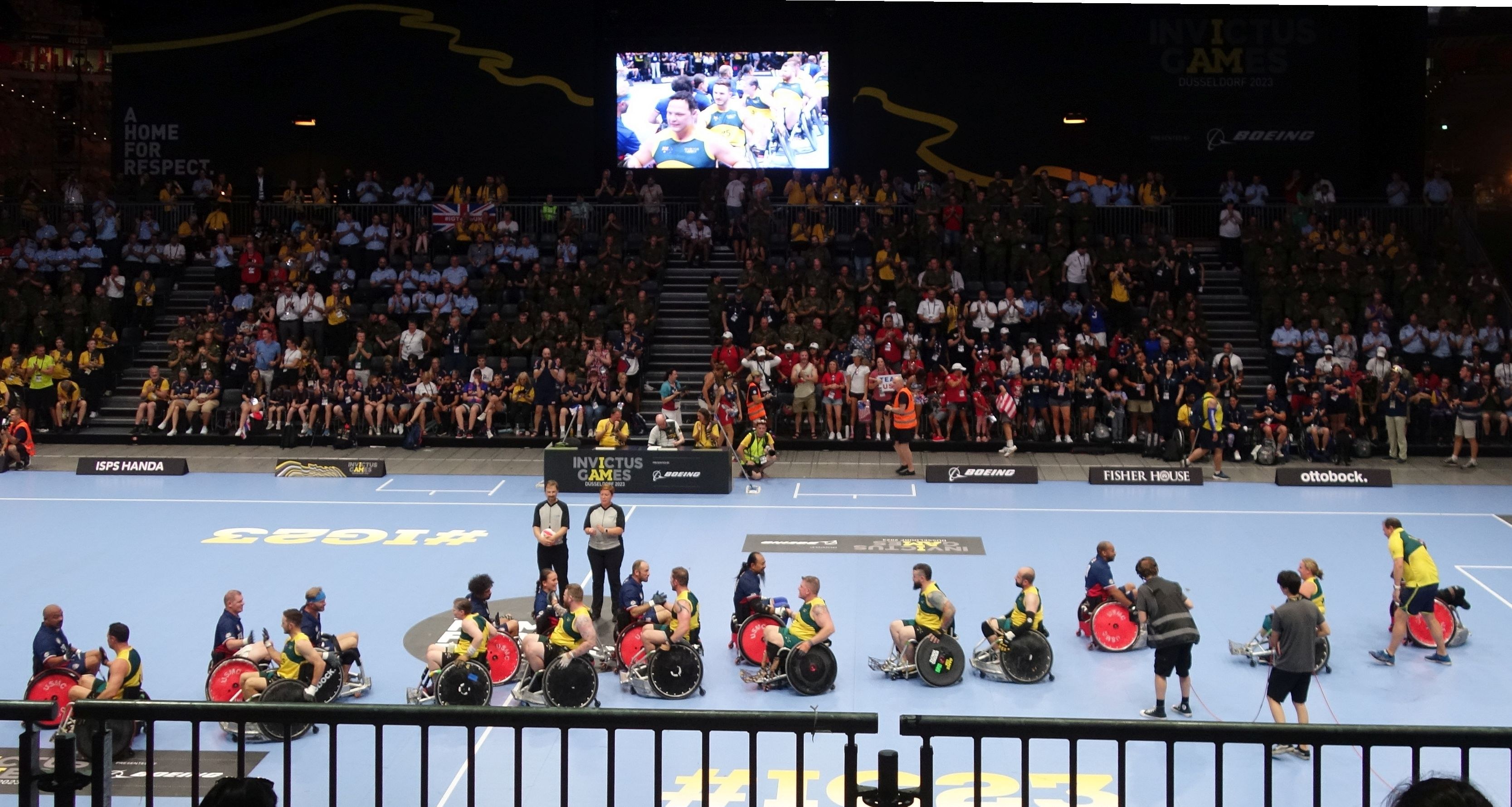 Eine Gruppe von Rollstuhlfahrern auf einem Platz bei den Invictus Games Düsseldorf 2020, umgeben von einer Menge, mit Treppen zu einem Bildschirm und Informationsplakaten im Hintergrund.