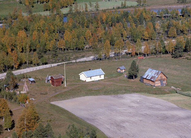 Eine Luftaufnahme eines Ranch mit verstreuten Gebäuden, umgeben von Feldern, Bäumen, Häusern, Pfählen, einer Straße, Gras und Wasser.
