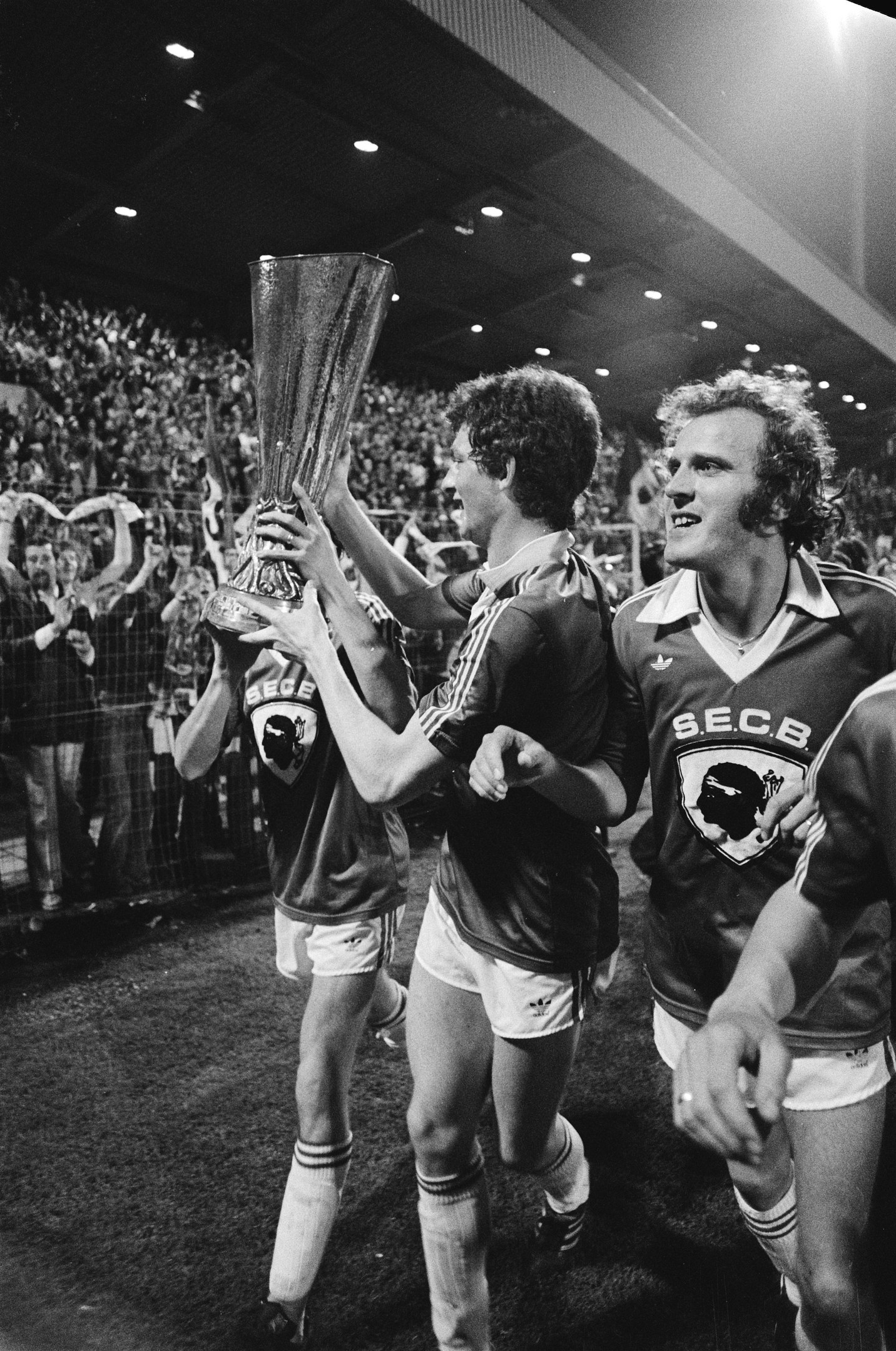 Schwarzes und weißes Bild von Fußballspielern, die auf einem Feld mit einem Pokal feiern, umgeben von einer Menge hinter einem Zaun unter Stadionbeleuchtung.