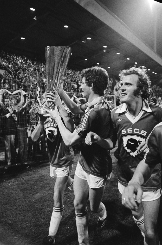 Schwarzes und weißes Bild von Fußballspielern, die auf einem Feld mit einem Pokal feiern, umgeben von einer Menge hinter einem Zaun unter Stadionbeleuchtung.