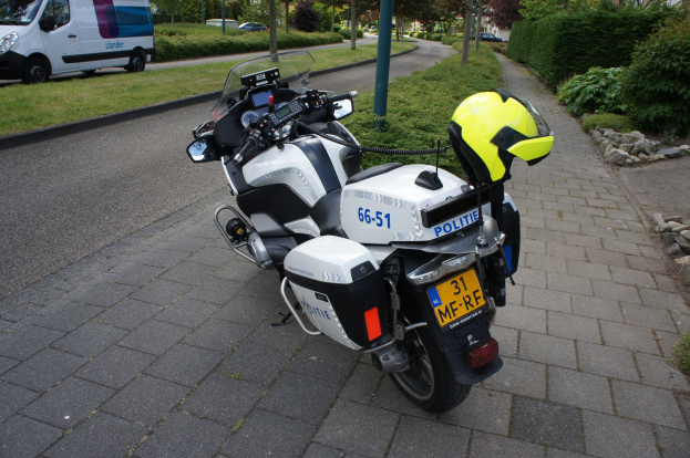 Polizeimotorrad am Straßenrand mit einem Helm darauf, umgeben von Bäumen, Pfählen, Gebäuden, Pflanzen, Gras, Steinen und anderen Fahrzeugen im Hintergrund.