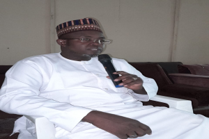 Ein Mann in einem weißen Gewand und einer Mütze sitzt in einem Stuhl und spricht in ein Mikrofon in einem Raum mit zwei Sofas und einer Wand, auf der der Satz "Ich bin kein Lehrer, ich bin ein Lehrer." steht.