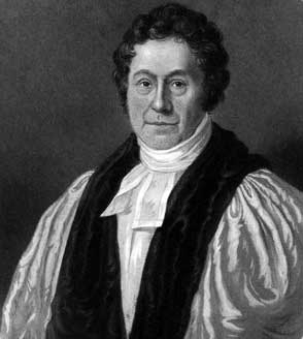 Ein Schwarz-Weiß-Foto eines Mannes in einem weißen Hemd und einem schwarzen Cape, mit ernstem Gesichtsausdruck, gefalteten Händen und konzentriertem Blick, der in einem schwach beleuchteten Raum mit spärlicher Einrichtung steht.