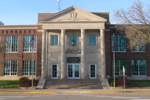 Kearney Junior High School-Gebäude mit Fenstern, Türen, Säulen, Stufen, einem Namensschild, Fahnen auf Stangen, Pflanzen, Gras, Bäumen und einer Straße davor, vor dem Himmel.