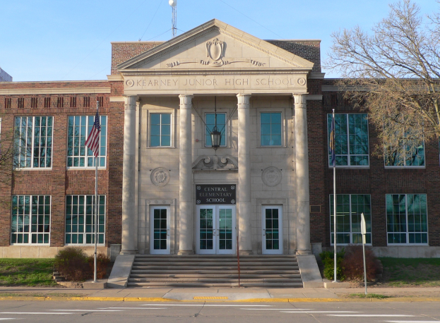 Kearney Junior High School-Gebäude mit Fenstern, Türen, Säulen, Stufen, einem Namensschild, Fahnen auf Stangen, Pflanzen, Gras, Bäumen und einer Straße davor, vor dem Himmel.
