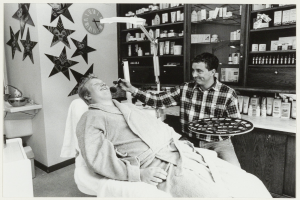 Ein Mann lächelt in einem Zahnarztstuhl, während ein anderer Mann neben ihm steht und einen Teller hält, mit einem Waschbecken, Sternen, einer Uhr und Regalen voller Flaschen im Hintergrund.