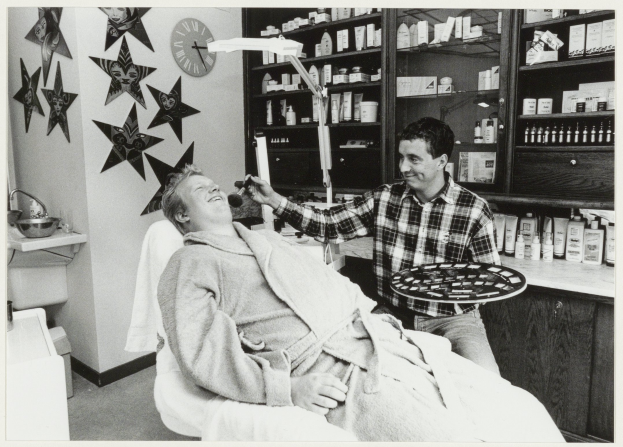 Ein Mann lächelt in einem Zahnarztstuhl, während ein anderer Mann neben ihm steht und einen Teller hält, mit einem Waschbecken, Sternen, einer Uhr und Regalen voller Flaschen im Hintergrund.