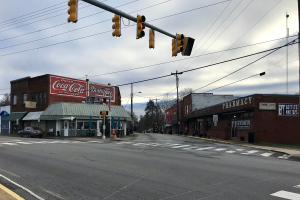Eine städtische Straßenkreuzung mit einem Verkehrslichtsignal, Fahrzeugen, Gebäuden mit Namensschildern, Strommasten mit Drähten, Bäumen und einem bewölkten Himmel.