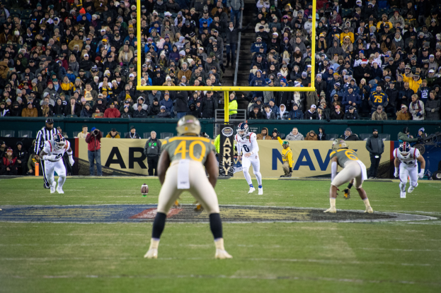 Fußballspieler in Sportkleidung und Helmen spielen ein Spiel in einem Stadion, während Zuschauer im Hintergrund zusehen, mit sichtbaren Schildern und Pfählen.