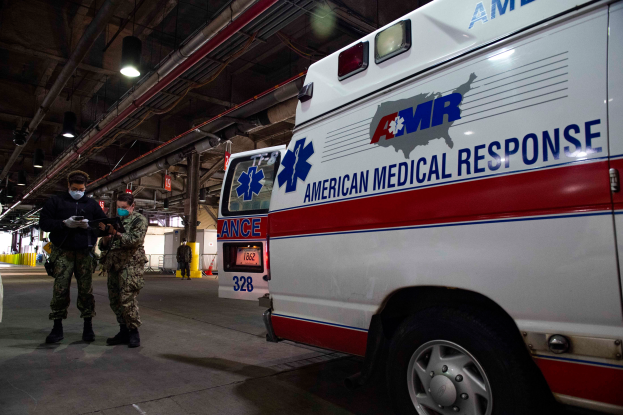 Ein Rettungswagen der American Medical Response (AMR) steht in einem Gebäude mit zwei maskierten Personen davor, einem Fahrzeug auf der linken Seite und mehreren Personen, Lichtern und Rohren im Hintergrund.